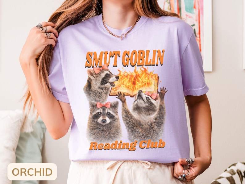 May include: Orchid-colored t-shirt with the text "SMUT GOBLIN Reading Club" in orange. The graphic features three raccoons, one with a book on fire. The raccoons wear pink bows. The shirt is a short-sleeved crew neck.