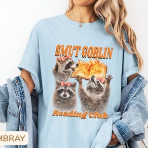 May include: Light blue t-shirt with the text "SMUT GOBLIN Reading Club" in orange. The graphic features three raccoons with pink bows, one holding a flaming book. The shirt has short sleeves and a relaxed fit.
