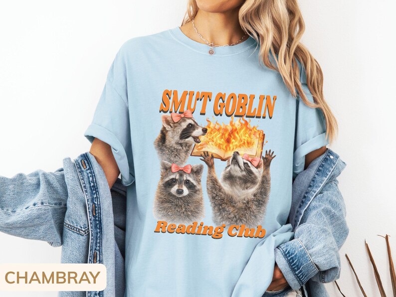 May include: Light blue t-shirt with the text "SMUT GOBLIN Reading Club" in orange. The graphic features three raccoons with pink bows, one holding a flaming book. The shirt has short sleeves and a relaxed fit.
