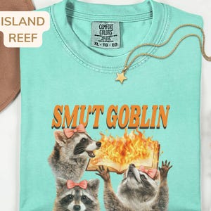 May include: A light turquoise t-shirt with the text "SMUT GOBLIN" in orange. The shirt features a graphic of three raccoons, two wearing pink bows, with one holding a burning book. A gold star pendant necklace is draped across the shirt.