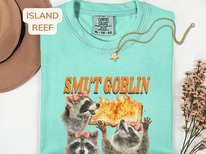 May include: A light turquoise t-shirt with the text "SMUT GOBLIN" in orange. The shirt features a graphic of three raccoons, two wearing pink bows, with one holding a burning book. A gold star pendant necklace is draped across the shirt.
