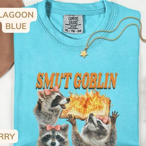 May include: Lagoon blue t-shirt with the text "SMUT GOBLIN" in orange. The shirt features a graphic of raccoons and a burning book. A gold star pendant necklace is draped on the shirt. The shirt is a Comfort Colors XL.