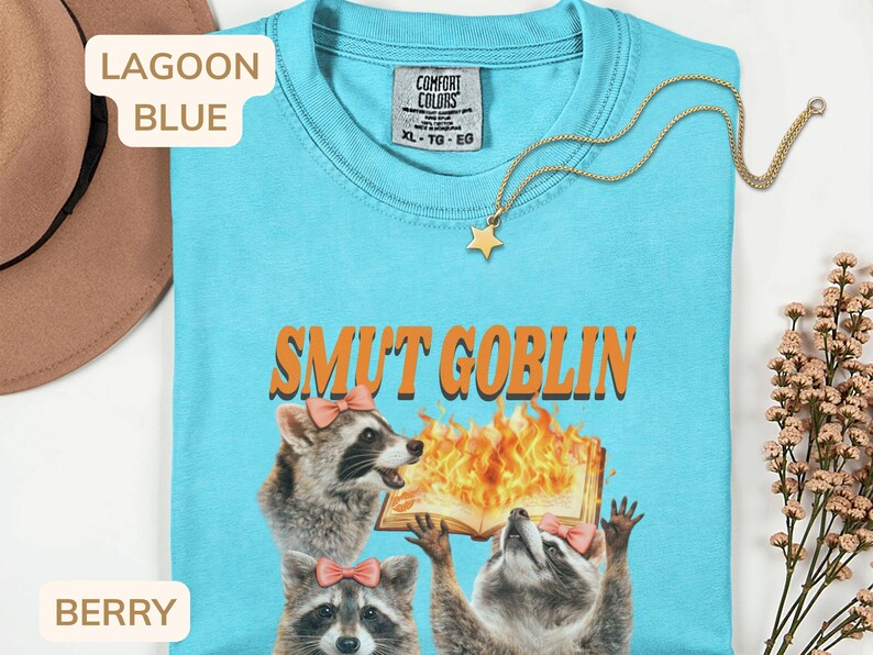 May include: Lagoon blue t-shirt with the text "SMUT GOBLIN" in orange. The shirt features a graphic of raccoons and a burning book. A gold star pendant necklace is draped on the shirt. The shirt is a Comfort Colors XL.
