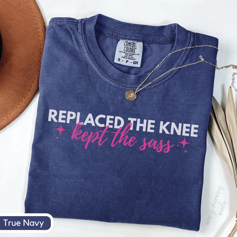 May include: A True Navy blue t-shirt with the text "REPLACED THE KNEE kept the sass" in white and pink. The shirt has a crew neck and short sleeves. A gold necklace with a pendant is draped across the shirt.