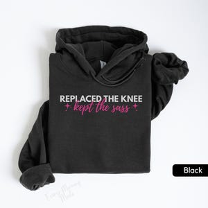 May include: Black hooded sweatshirt with the text "REPLACED THE KNEE kept the sass" in white and pink. The sweatshirt has a drawstring hood and long sleeves. The word "Black" is in a black rectangle in the bottom right corner.