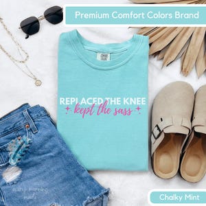 May include: A Chalky Mint Comfort Colors t-shirt with the text "REPLACED THE KNEE + kept the sass +". The shirt is displayed with denim shorts, sunglasses, a necklace, and sandals.