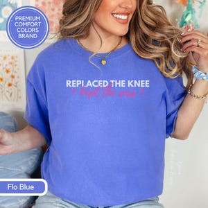 May include: Flo Blue t-shirt with the text "REPLACED THE KNEE * kept the sass *" in white and pink. The shirt has a crew neck and short sleeves. The brand logo is in the upper left corner. The shirt is a comfort colors brand.