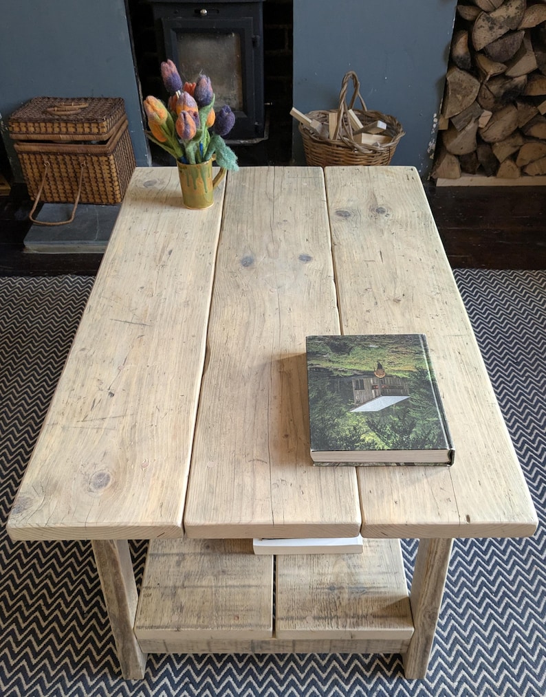 May include: A rectangular coffee table constructed from light-toned reclaimed wood planks with a lower shelf. The table surface features a book with a green forest cover and a yellow mug containing a bouquet of felted orange, purple, and blue tulips.
