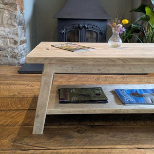 May include: A rectangular light-colored wooden coffee table with a lower shelf. The top surface holds a magazine and a small glass vase with dried flowers. The lower shelf contains two magazines, one titled Gamewise and one titled Shooting.