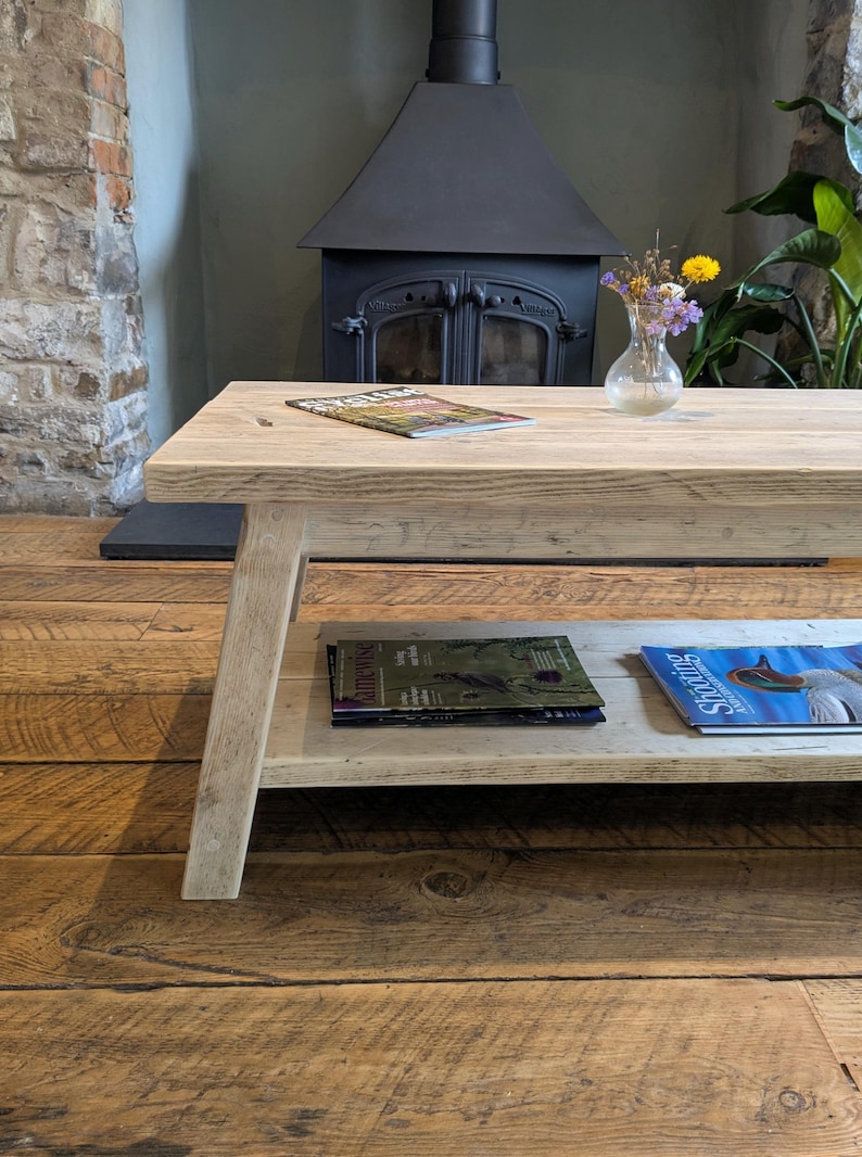 May include: A rectangular light-colored wooden coffee table with a lower shelf. The top surface holds a magazine and a small glass vase with dried flowers. The lower shelf contains two magazines, one titled Gamewise and one titled Shooting.