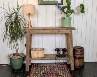 May include: A rustic wooden console table with a woven rug, potted plants, and a framed poster. The table holds a lamp, a basket, and a vintage telephone. The rug has a geometric pattern with various colors.