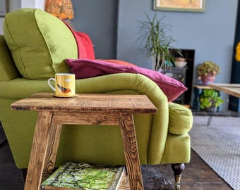 May include: A rustic wooden side table with a shelf. The table is stained a dark brown and has a distressed finish. A book titled "Simple Trees" and a mug with a bird design are on the table. The table is next to a green sofa with red and purple pillows.