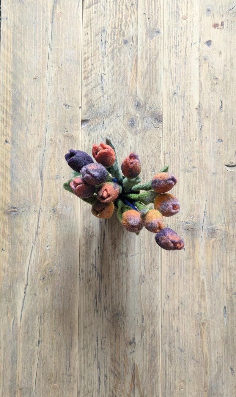 May include: A bouquet of needle-felted wool tulips in shades of deep purple, burnt orange, and muted yellow. Each flower features a rounded, closed bud shape with soft, textured petals and green felt stems bundled together in a tight cluster.