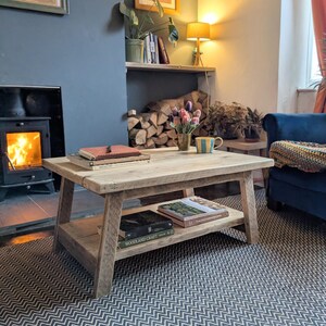 May include: A rectangular light-toned wooden coffee table with a lower shelf. The table features a thick, plank-style top and four sturdy, slightly angled legs. Books are stacked on both the top surface and the lower shelf of the table.