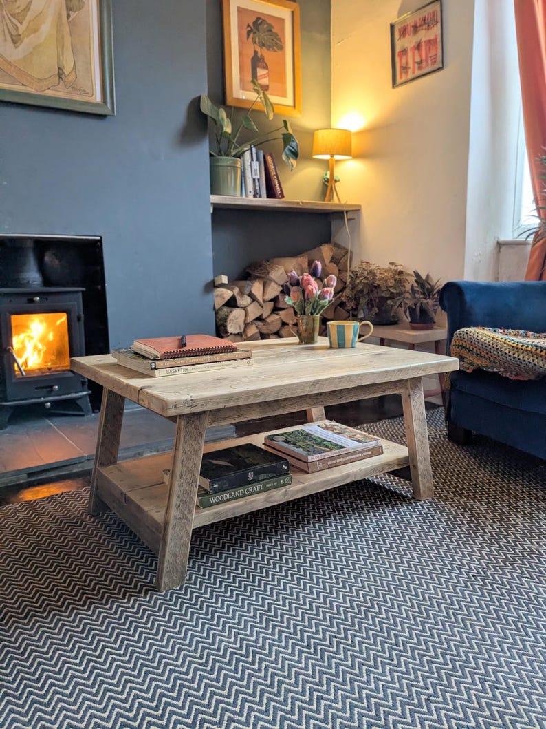 May include: A rectangular light-toned wooden coffee table with a lower shelf. The table features a thick, plank-style top and four sturdy, slightly angled legs. Books are stacked on both the top surface and the lower shelf of the table.