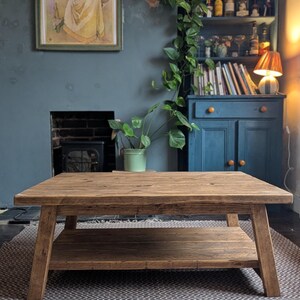 May include: A rectangular coffee table constructed from medium-brown wood with a visible grain pattern. The table features a flat top surface supported by four angled legs and includes a lower shelf positioned between the legs for additional storage.