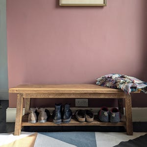 May include: A wooden bench with a lower shelf, displaying shoes. A framed artwork hangs on a mauve wall. A colourful blanket rests on the bench. The rug below has a geometric pattern.