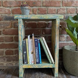 May include: A small, rustic wooden side table with a distressed teal and yellow finish. The table holds several books, including titles like "Mushrooms" and "Wild Swim." A small potted succulent sits on top. The background features a brick wall.