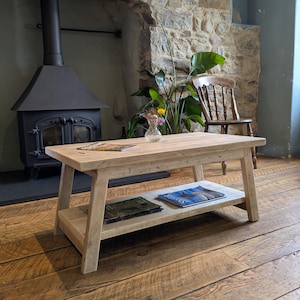 May include: A rectangular light-colored wooden coffee table with a smooth top surface and a lower shelf. The table features four angled legs and a natural wood grain finish. Magazines are placed on the top surface and the lower shelf.