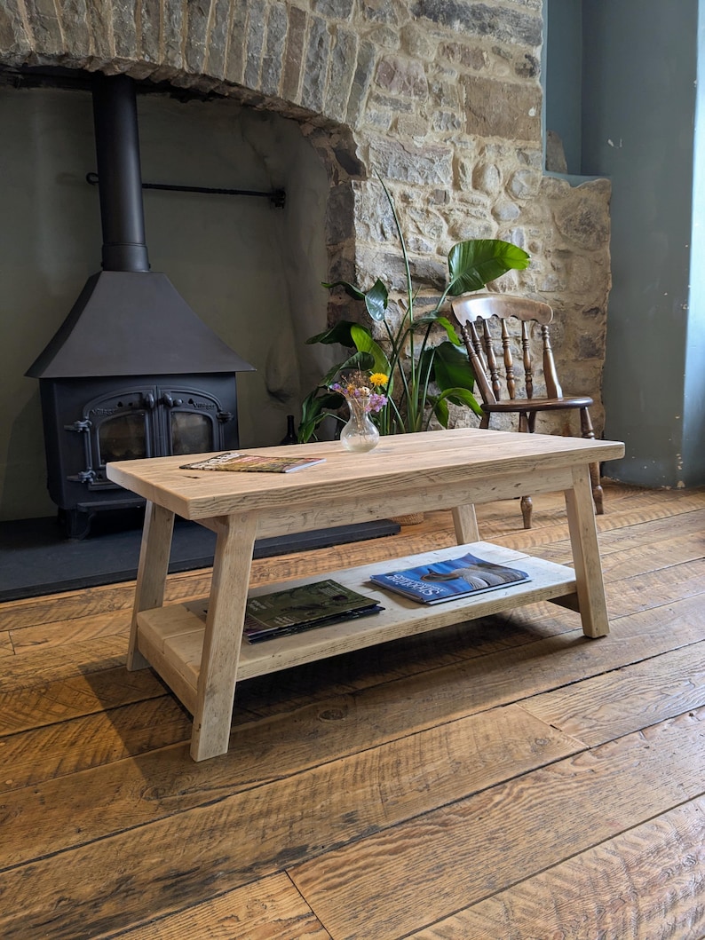 May include: A rectangular light-colored wooden coffee table with a smooth top surface and a lower shelf. The table features four angled legs and a natural wood grain finish. Magazines are placed on the top surface and the lower shelf.