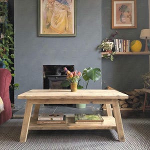 May include: A rectangular light-toned wooden coffee table with a lower shelf. The table features four angled legs and a natural wood grain finish. Two books are placed on the lower shelf, and a yellow vase with colorful tulips sits on top.