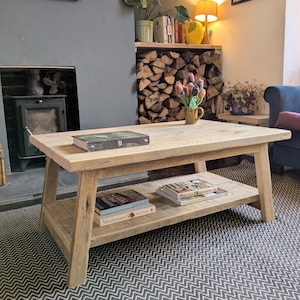 May include: A rectangular coffee table constructed from light-toned reclaimed wood featuring a thick tabletop and a lower storage shelf. The table has four angled legs and supports several books on both the top surface and the bottom shelf.