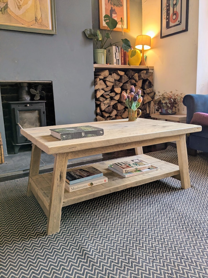 May include: A rectangular coffee table constructed from light-toned reclaimed wood featuring a thick tabletop and a lower storage shelf. The table has four angled legs and supports several books on both the top surface and the bottom shelf.