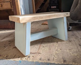 Puede incluir: Un banco de madera con un asiento rectangular grueso de tono natural y veta visible. La base consta de dos patas pintadas en azul claro conectadas por una viga de soporte horizontal, creando una estructura resistente para este mueble pequeño.