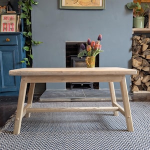 May include: A light oak bench with angled legs. A small vase of colourful flowers sits on the bench. The bench is placed on a blue and white patterned rug. The background includes a fireplace and artwork.