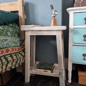 May include: A light-toned wooden side table with a lower shelf. A small, textured vase with feathers sits on top, next to a book. A stack of books is on the lower shelf. The table is next to a bed and a painted cabinet.