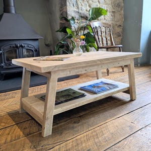 May include: A rectangular light-toned wooden coffee table featuring a lower shelf for storage. The table has four angled legs and a natural wood grain finish. Two magazines are placed on the lower shelf, and one magazine sits on the top surface.