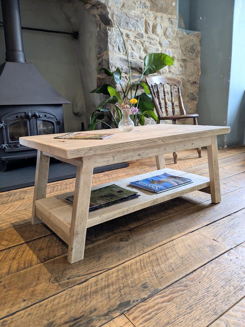 May include: A rectangular light-toned wooden coffee table featuring a lower shelf for storage. The table has four angled legs and a natural wood grain finish. Two magazines are placed on the lower shelf, and one magazine sits on the top surface.