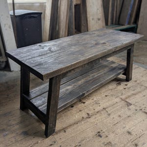 May include: A rectangular wooden bench featuring a dark charcoal-stained finish with visible wood grain. The bench is constructed with four sturdy legs and includes a lower shelf positioned between the legs, providing an additional flat surface.