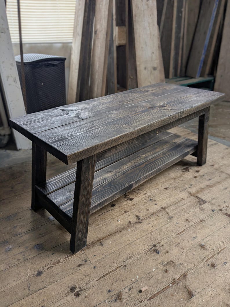 May include: A rectangular wooden bench featuring a dark charcoal-stained finish with visible wood grain. The bench is constructed with four sturdy legs and includes a lower shelf positioned between the legs, providing an additional flat surface.