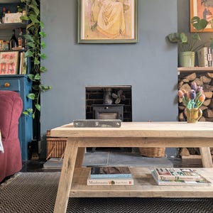 May include: A rectangular light-toned wooden coffee table with a lower shelf. The table features angled legs and a natural wood grain finish. Several books are stacked on the lower shelf, and one book rests on the top surface of the table.