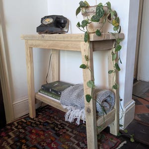 May include: A light-coloured wooden side table with a black rotary phone, a potted plant, books, and a rolled blanket. The table has two shelves and is placed on a patterned rug. The plant has green and yellow leaves.