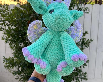May include: A crocheted green dragon plush toy with purple accents. The dragon has a long tail, wings, and a friendly face.