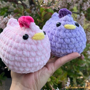 May include: Two crocheted plush chicks, one pink with a pink crest and one purple with a purple crest. Both chicks have black eyes and yellow beaks.