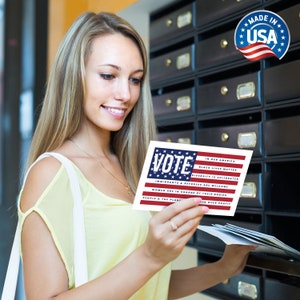 May include: A woman holds a card with an American flag design and the word "VOTE" in bold letters. The card also includes the text "In our America, Black Lives Matter, Diversity is Celebrated, Immigrants & Refugees are Welcome, Women are in Charge of their Bodies, People & the Planet over Profit."