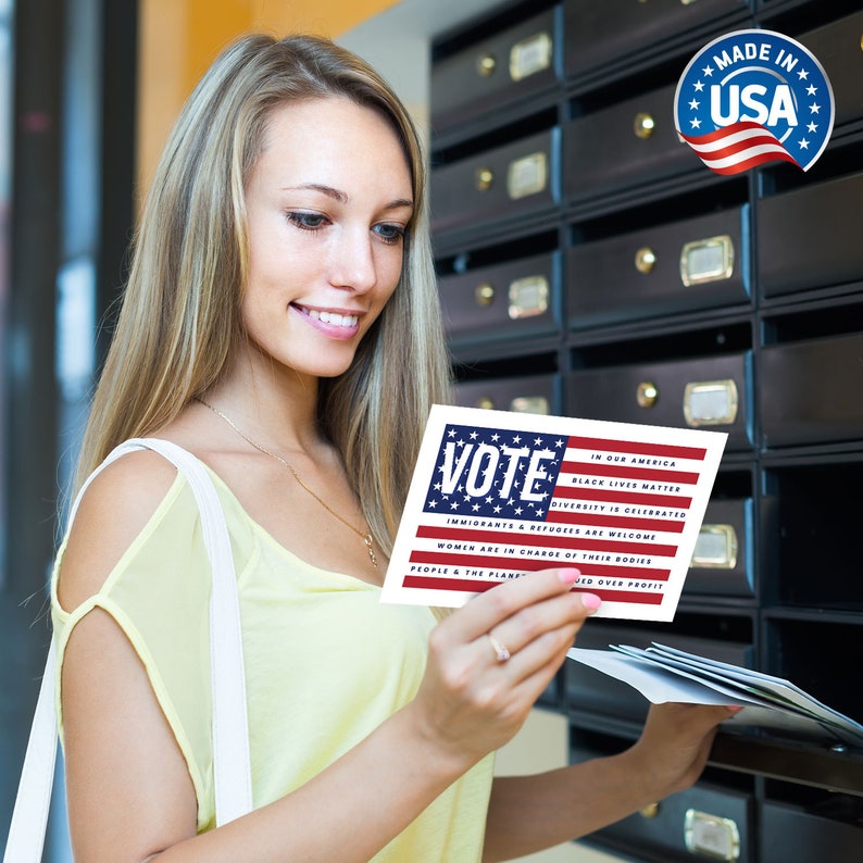 May include: A woman holds a card with an American flag design and the word "VOTE" in bold letters. The card also includes the text "In our America, Black Lives Matter, Diversity is Celebrated, Immigrants & Refugees are Welcome, Women are in Charge of their Bodies, People & the Planet over Profit."
