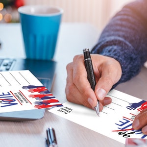 Może przedstawiać: Dwie białe kartki pocztowe z niebieskimi i czerwonymi odciskami dłoni i tekstem "Vote" oraz "Moc jest w twoich rękach!" Tekst "Umieść znaczek tutaj" znajduje się na prawej kartce. Osoba pisze na kartce czarnym długopisem.
