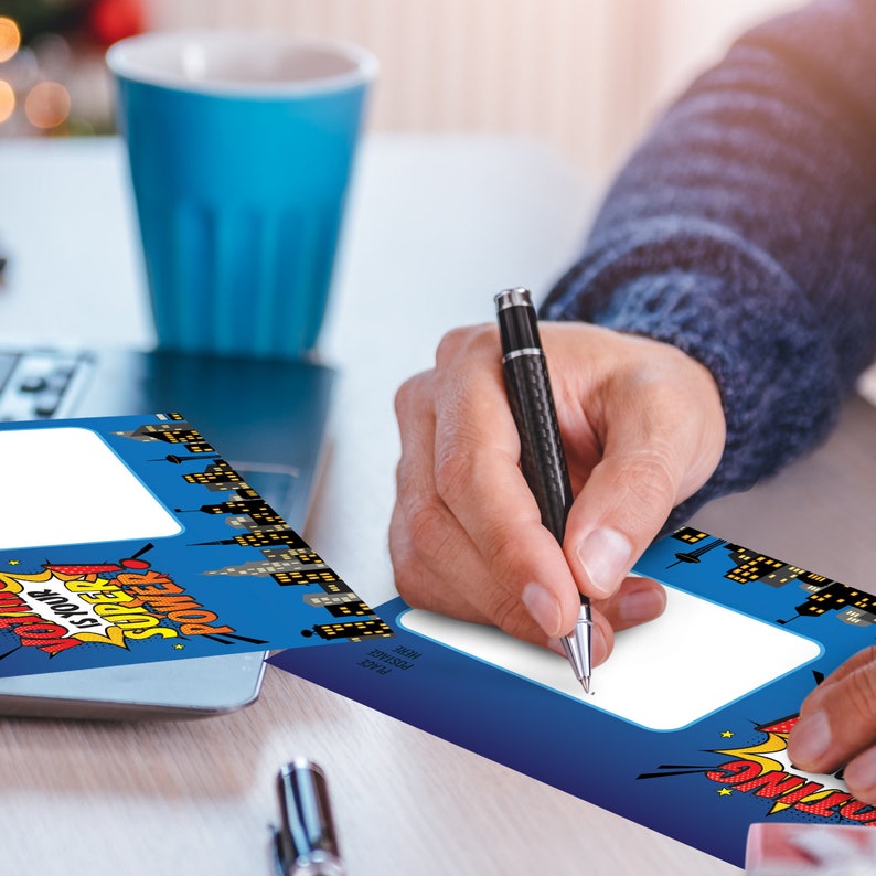 May include: Two blue and white greeting cards with a cityscape and comic book style design. The text on the card reads "Power is your Superpower". A person is writing on one of the cards with a black pen.