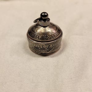 May include: A silver half dollar coin has been transformed into a small, round, hinged container. The coin's original design is visible on the top and bottom of the container. The container has a small, black metal loop on top.