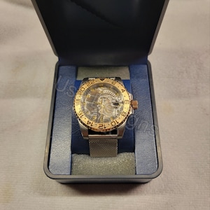 May include: A wristwatch with a silver-tone mesh band and a gold-tone rotating bezel featuring black numerals. The watch face displays a detailed embossed profile of a figure, gold-tone hands, and the text IN GOD WE TRUST in a dark gray finish.