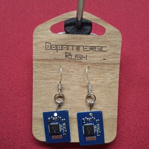 May include: A pair of unique earrings featuring small, blue circuit boards with silver-tone hooks. The earrings are attached to a wooden display card with the text "Dopaminergic Rush" in a modern font. The background is a solid red.