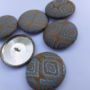 May include: A collection of six large, round buttons. The buttons have a textured, patterned design in shades of gold and blue. One button is turned over, revealing a silver metal back with a loop for sewing.