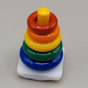 以下が含まれることがあります： 白いベースのミニチュア木製スタッキングおもちゃ。青、緑、黄、オレンジ、赤の5色のリングと黄色のペグが付いています。小さなお子様向けに設計されています。