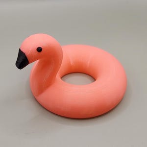 Puede incluir: Un flotador de piscina con forma de flamenco rosa. El flamenco tiene un pico y un ojo negros, y un cuerpo circular. El flotador está hecho de un material liso y sólido. El flamenco es un artículo decorativo y divertido.