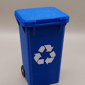 May include: A miniature blue recycling bin with a hinged lid and a white recycling symbol printed on the front. The bin features a textured surface finish and includes small black wheels attached to the lower sides of the rectangular container.