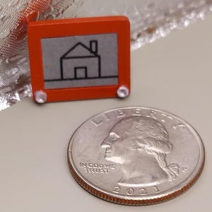 May include: Miniature orange Etch-A-Sketch toy with a gray screen featuring a house drawing. The toy is next to a U.S. quarter for size comparison. The Etch-A-Sketch has two white knobs and a red handle.